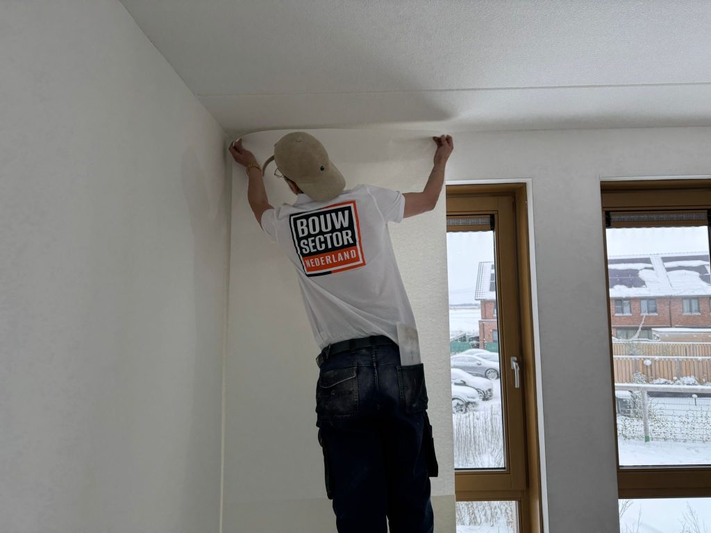 Vakman van Bouwsector Nederland zet een baan vlies bovenaan tegen het plafond vast naast twee houten ramen