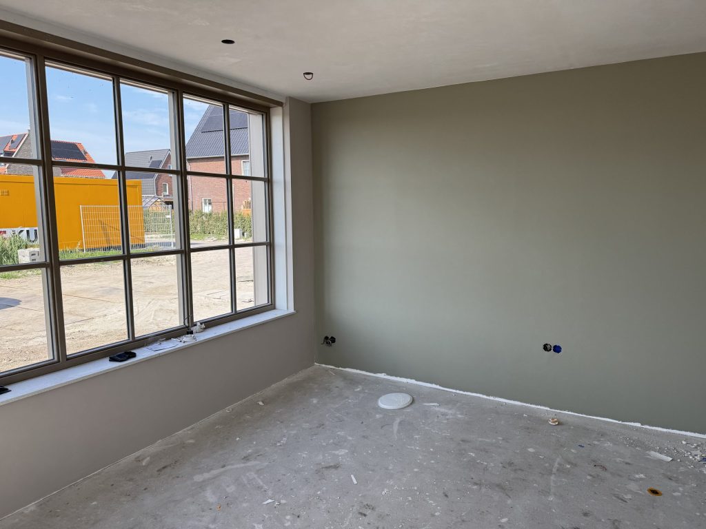 Nieuwbouwkamer met een egaal gesausde grijsgroene wand en een groot raam op een bouwterrein