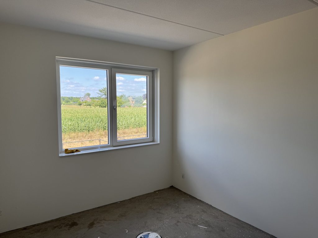 Lege nieuwbouwkamer met gladde, lichte wanden, een betonvloer en een raam met uitzicht op een maisveld