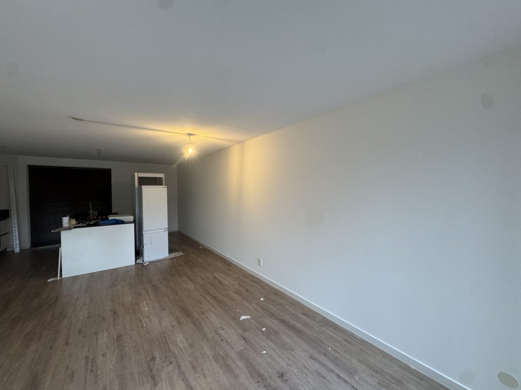 Open woonkamer van een appartement met een lange, strak gesausde witte wand en een keukenblok achterin