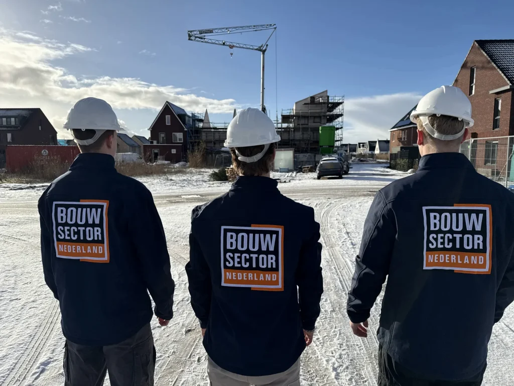 Drie vakmensen van Bouwsector Nederland met witte helmen en donkerblauwe jassen met logo kijken naar een nieuwbouwwijk in de sneeuw